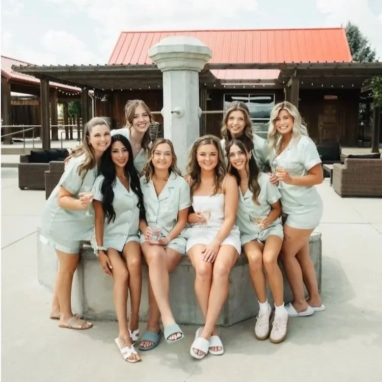 Six young bridesmaids sitting on a stone fountain, wearing pajamas, with the bride in a white satin pajama top and pants, all smiling and hugging, outdoor setting with a cloudy sky and mountains in the background.