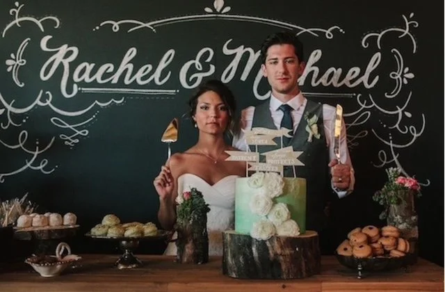 Couple at a wedding reception with a decorated table and a chalkboard background with the names Rachel and Michael.