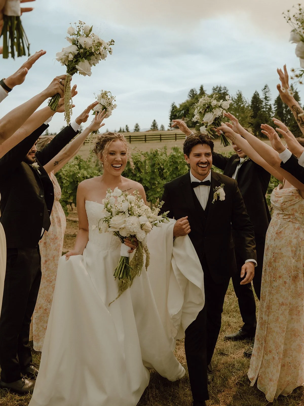 The bride and groom walk together, smiling, surrounded by friends and family throwing bouquets in a celebratory outdoor wedding scene with vineyards and trees in the background.