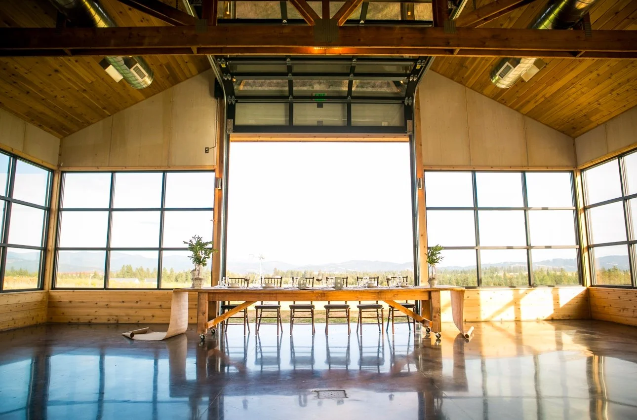 Large modern, elegant and rustic wedding space with high warm wood ceiling, wood trusses, dark polished concrete floors,  large windows and large garage door with mountain and vineyard views. Farm table with Chiavari chairs and 2 potted plants.