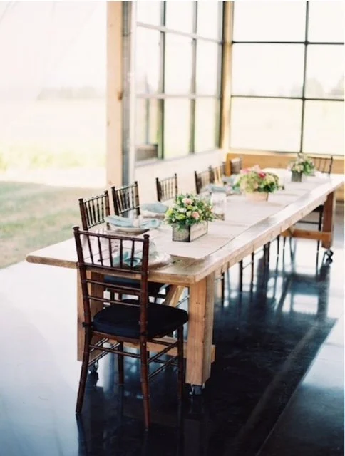 A long wooden farm table set with floral centerpieces, plates, and napkins in a bright room with large windows and wooden chairs.