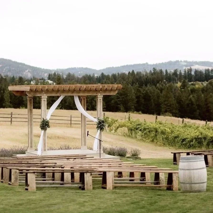 An outdoor wedding ceremony setup with an empty wooden stage, white drapes, and floral decorations, set against a scenic backdrop of trees, vineyards and mountains.