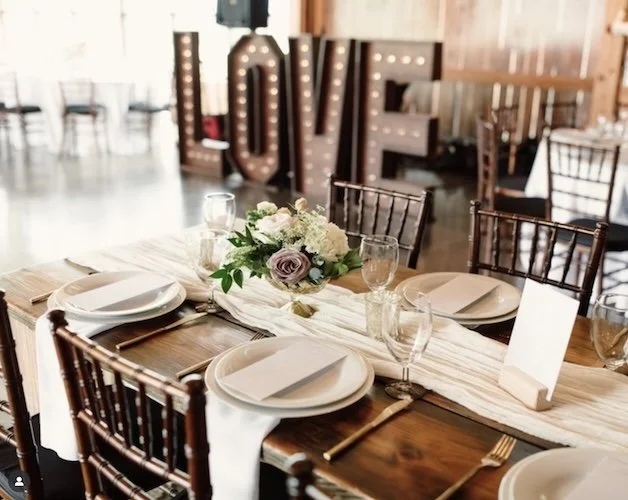 Elegant dining table set with white plates, glasses, cutlery, a floral centerpiece, and a light-colored table runner, with illuminated decorative "LOVE" sign in the background.