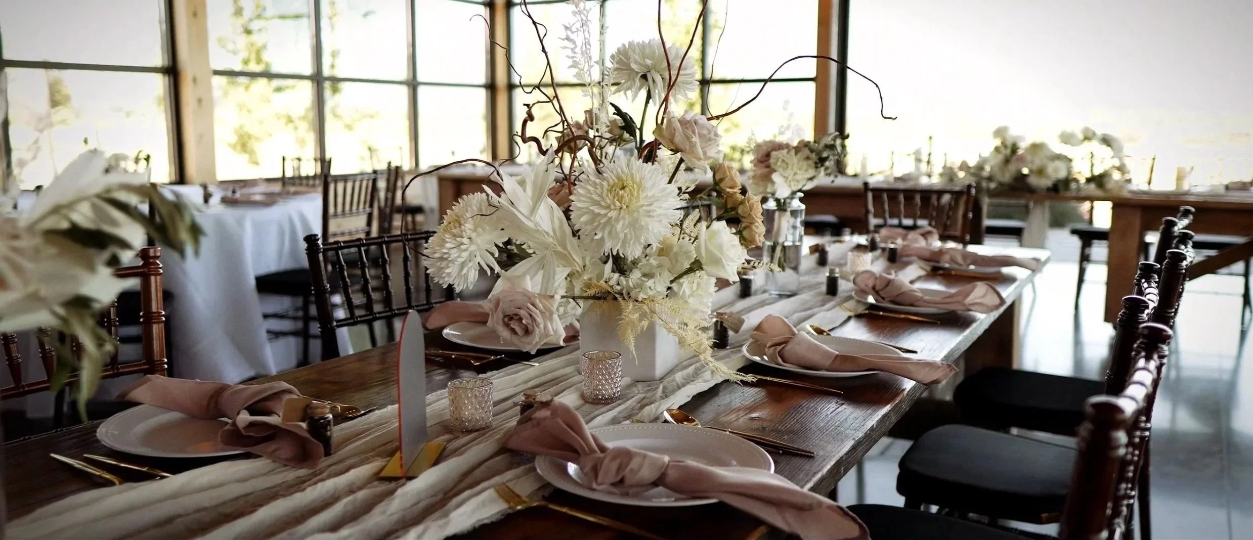 A long wooden dining table decorated for a wedding with a large white floral centerpiece, pink and beige napkins, gold cutlery, and small candle holders, set in a bright barn with large windows.