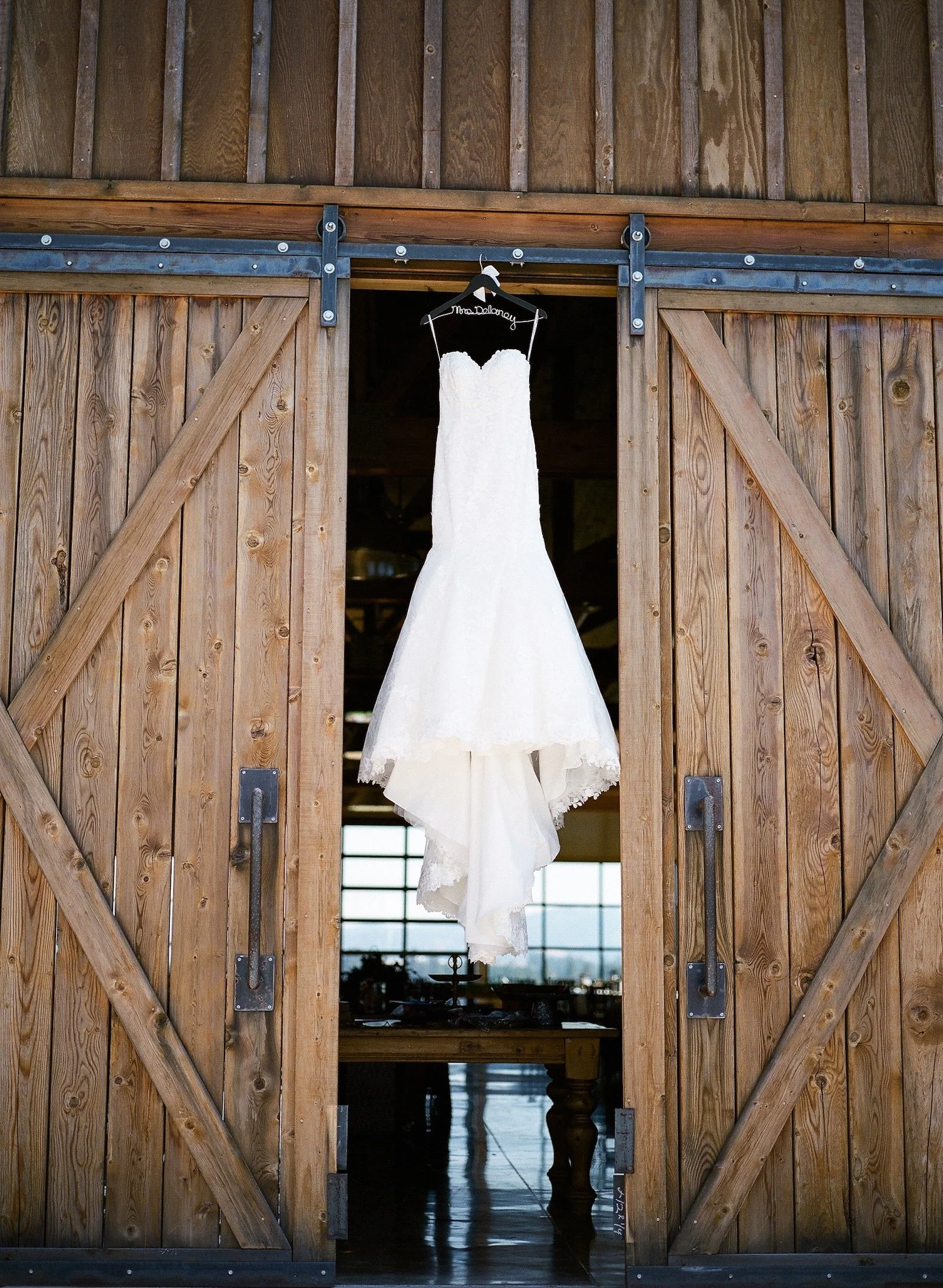 A white wedding dress hanging on a black hanger with a name wire that reads 'Mrs. Doloney', hanging from a sliding barn door in a rustic venue.