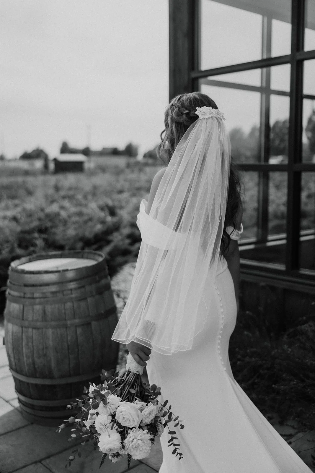 A bride in a white wedding dress with buttons down the back, holding a bouquet of flowers, standing outdoors near a barn with large glass windows and a wine barrel, with a vineyard in the background.