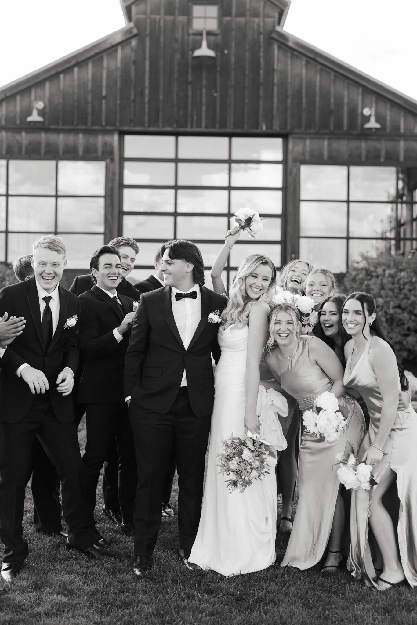 Black and white photo of a wedding party standing outdoors in front of a large barn with glass windows. The group, consisting of men and women in formal attire, is smiling and laughing. The bride in a white dress is holding a bouquet, and the groom i