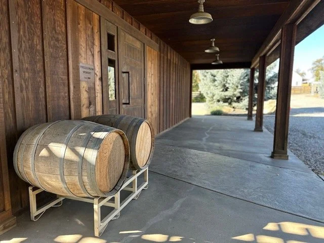 Two wooden wine barrels placed on a metal stand against a tasting room barn wall in an outdoor covered area, with trees and open space visible in the background.