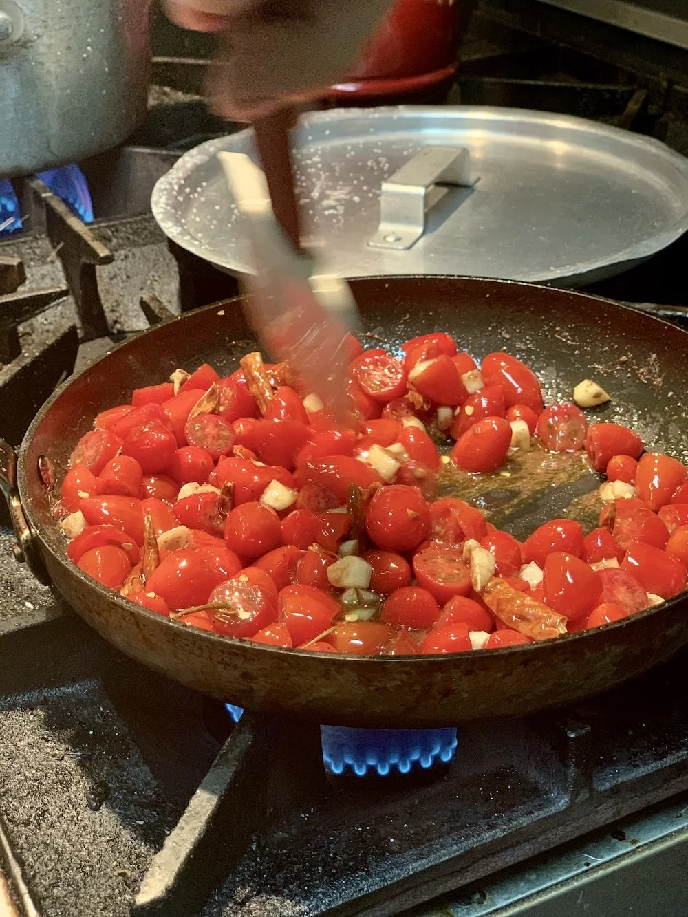 Chopped cherry tomatoes, garlic, and herbs being sautéed in a black skillet on a gas stove.