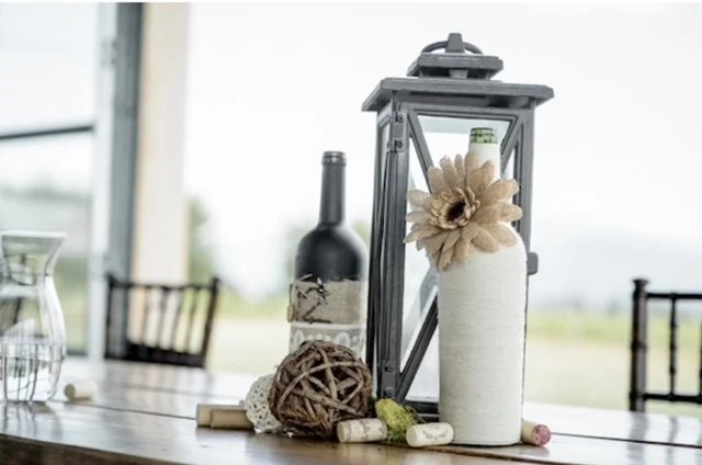 Decorative table centerpiece with a tall white vase with flowers, a wine bottle, a wooden ball, and colorful chalks on a wooden table.