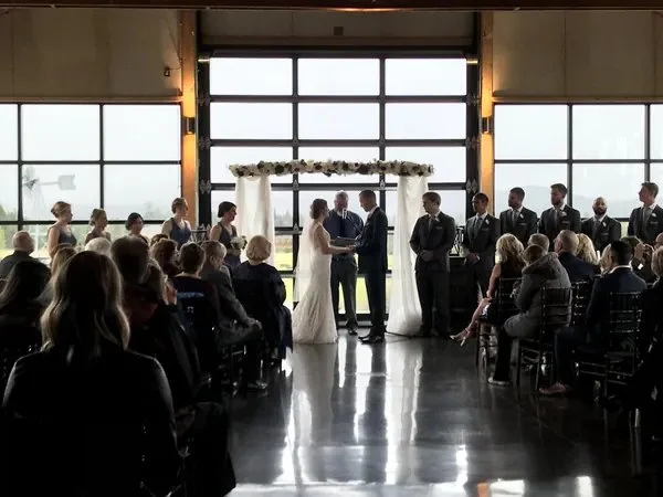 Bride and groom exchanging vows beneath a floral wedding arch in a modern rustic barn venue surrounded by wedding guests and attendants.