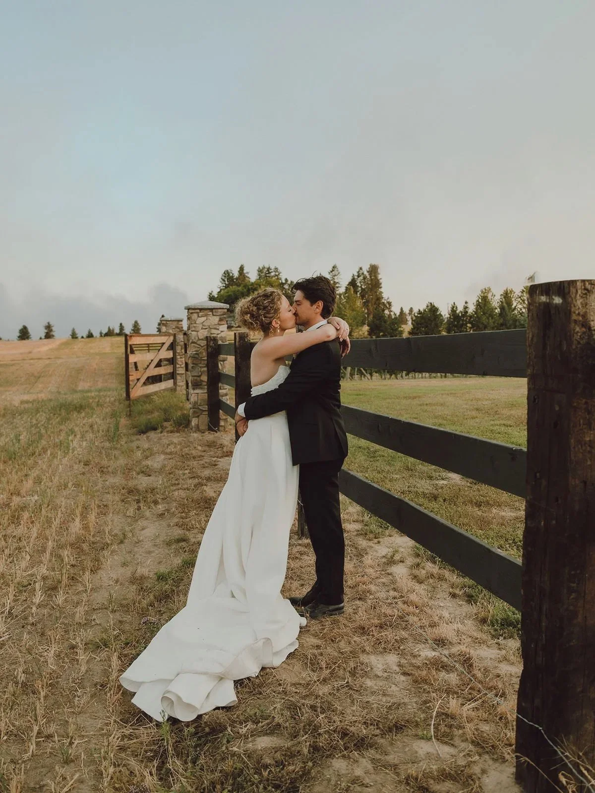 A bride and groom embrace and kiss outdoors in a rural setting with a wooden fence, grassy fields, and trees in the background.