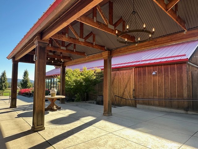 Covered atrium with wooden beams, a chandelier, and a concrete floor, adjacent to a building with a red metal roof and a garden with trees and shrubs.