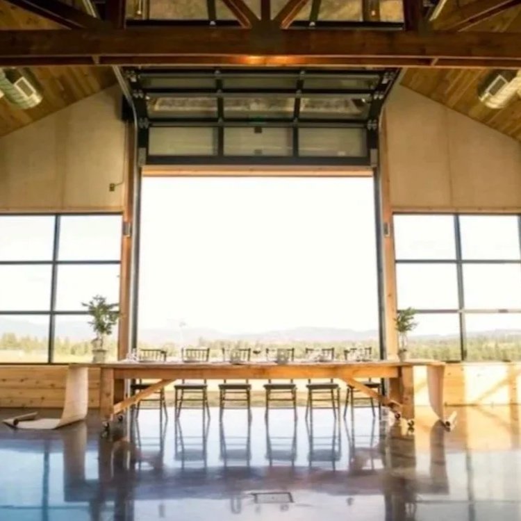 Interior of a spacious room with large windows, a wooden ceiling, and a long wooden table set with plates, glasses, and two potted plants at each end, overlooking a scenic outdoor landscape.
