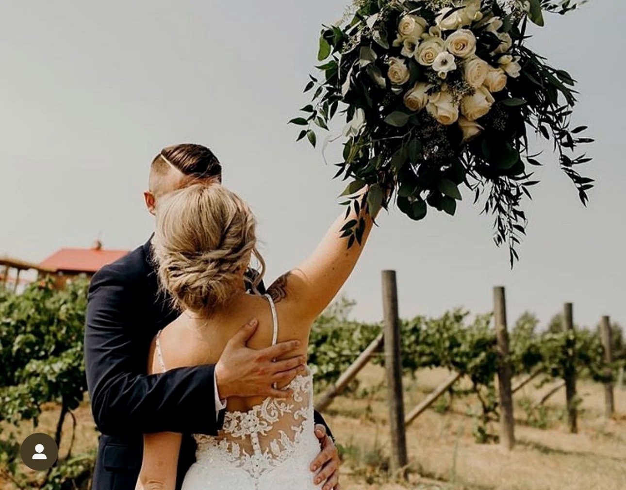 Bride and groom embrace in a vineyard during a wedding, with the bride holding a large bouquet of white flowers and greenery.