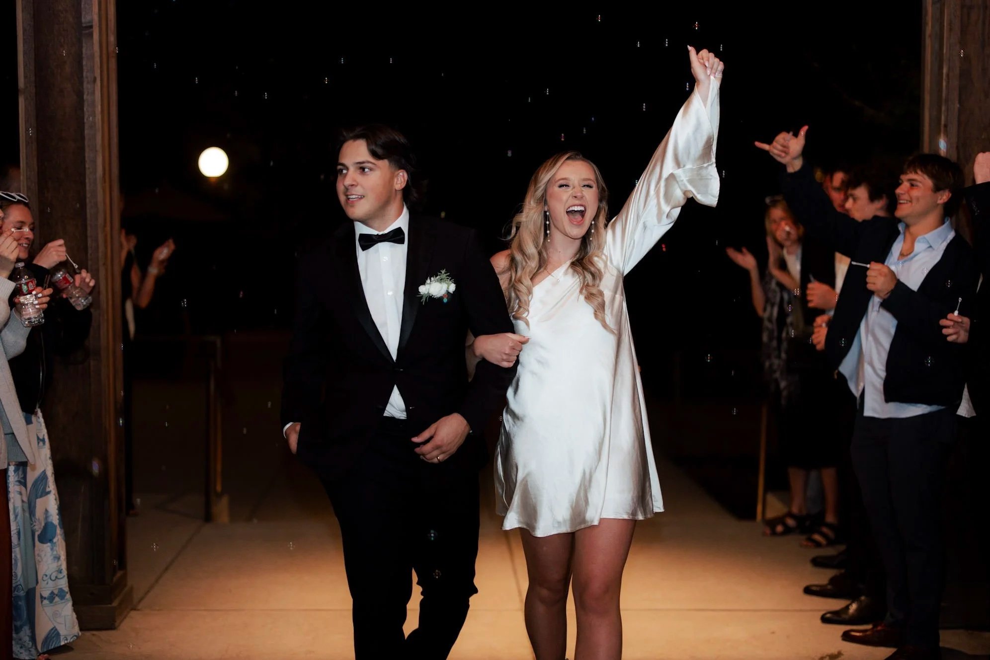 A couple walking arm-in-arm at a wedding reception send-off, surrounded by smiling guests. The woman is wearing a white dress and raising her arm, while the man is dressed in a black tuxedo with a bowtie.