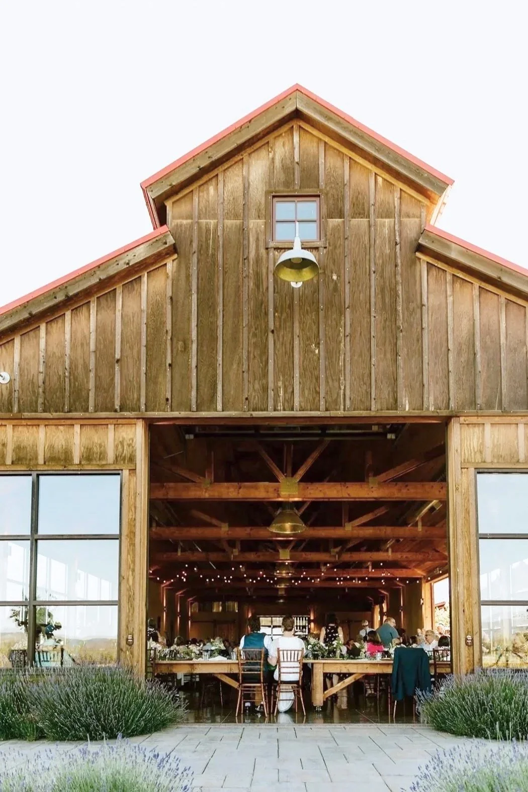 Rustic barn converted into a wedding reception venue with guests seated at long tables inside, decorated with flowers, with string lights hanging from the ceiling.