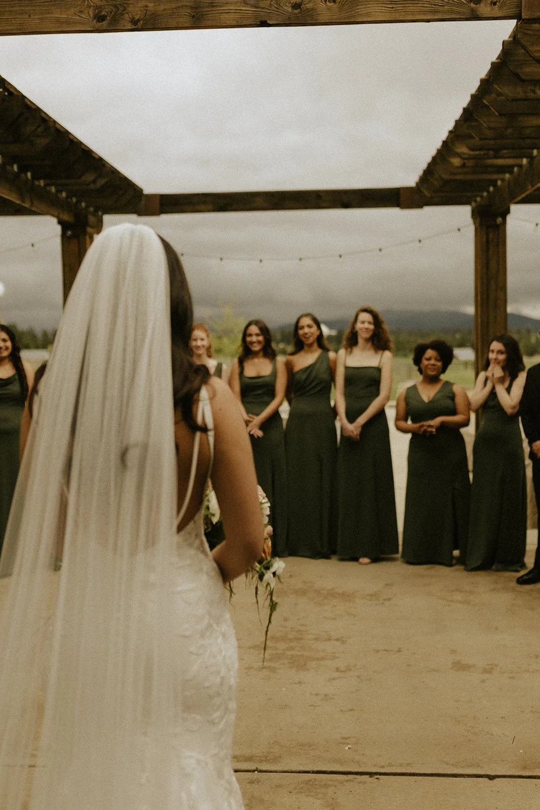 Bride holding a bouquet faces bridesmaids in dark green dresses beneath a wooden outdoor pergola.