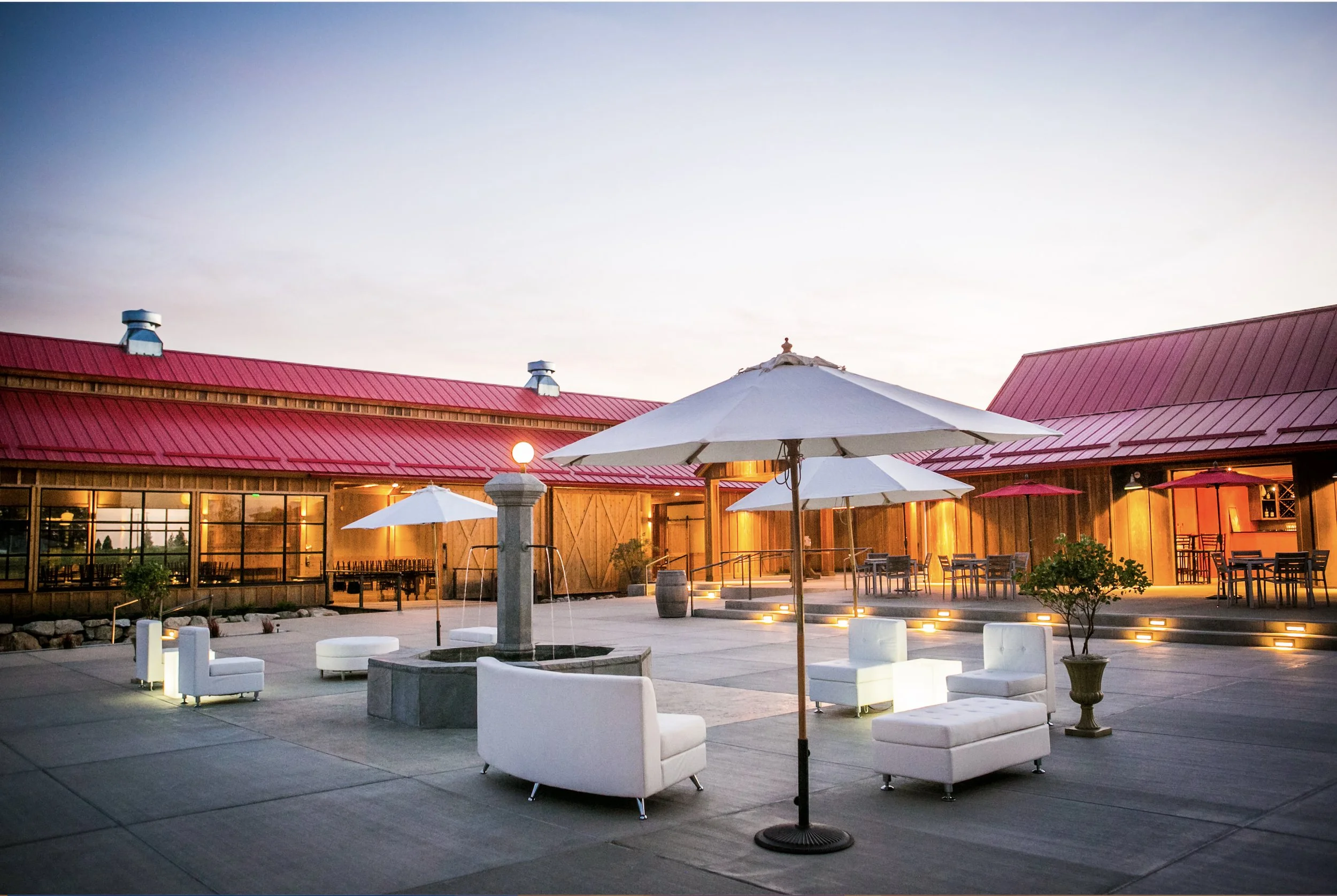 Outdoor courtyard with white lounge chairs, tables with umbrellas, and a building with red roof and large windows at dusk.