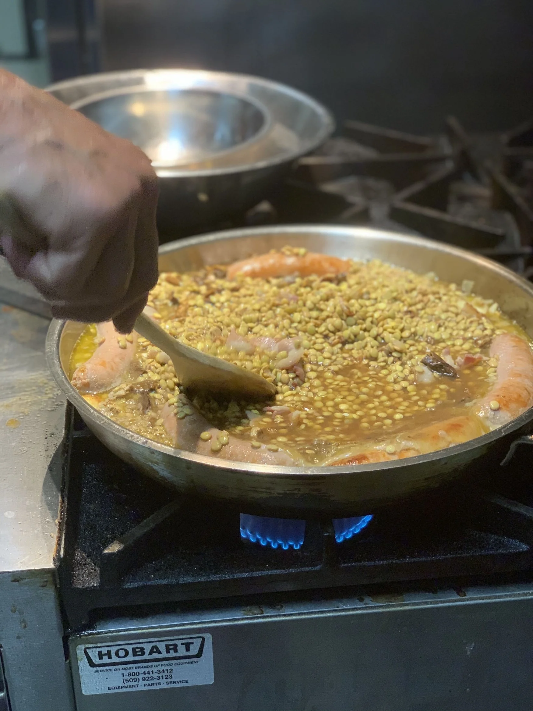 Cooking a risotto dish with saffron and sausages in a sauté pan on a stove with blue flames, while the person cooking stirs with a wooden spoon.