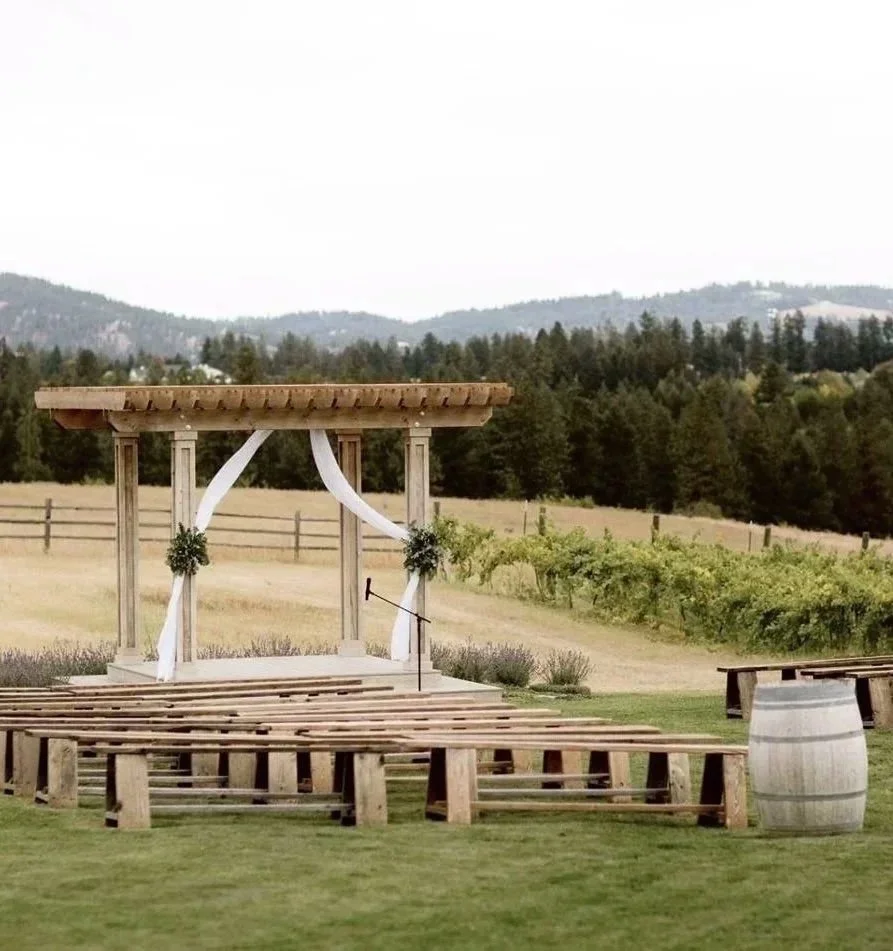 An outdoor wooden step-up wedding arbor draped with white fabric and greenery, set on a grassy field with ceremony benches and a wine barrel nearby, with a background of  vineyards, rolling farmland and trees.
