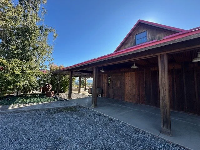 A wooden barn with a red roof, covered porch, and a gravel driveway, set against a clear blue sky.