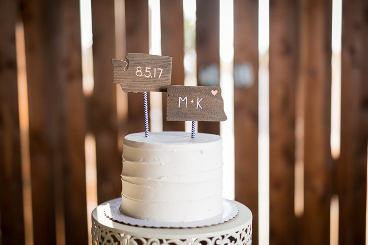 A white frosted wedding cake with two wooden signs on top, one with the date 8.5.17 and the other with the initials M+K, in front of a barn door.