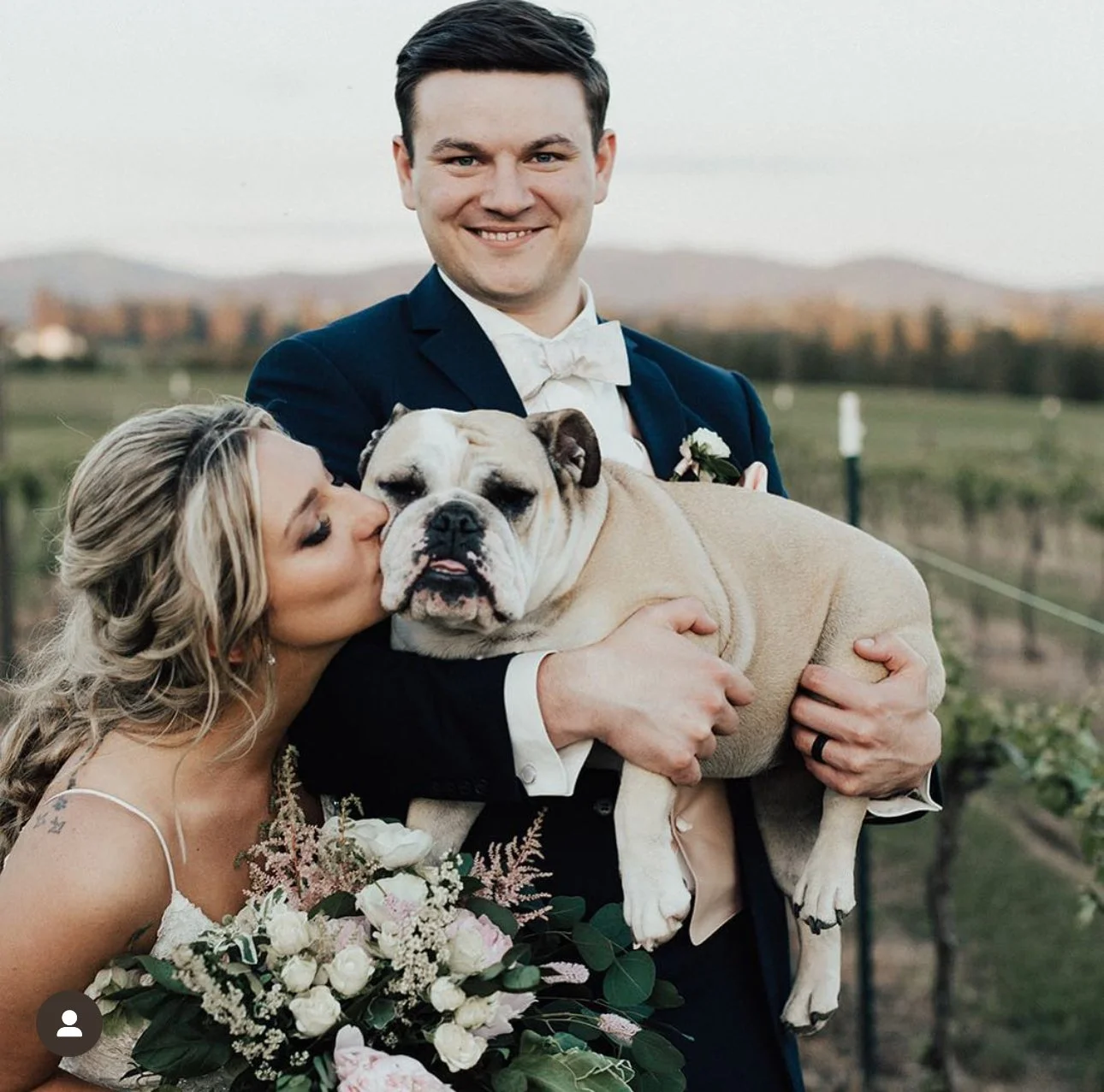 A newlywed couple with a bulldog at an outdoor spring wedding, the woman kisses the dog while holding a bouquet, the man holds the dog and smiles at the camera, in a vineyard setting.