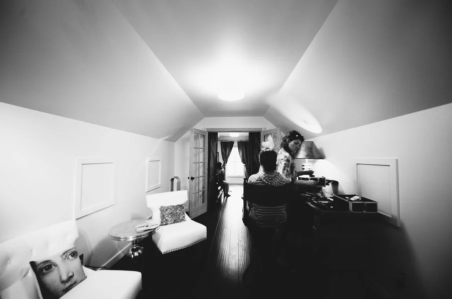 A black and white photo of a cozy bridal suite with two people near a desk, a window with curtains, and a sitting area with a pillow on the chair.