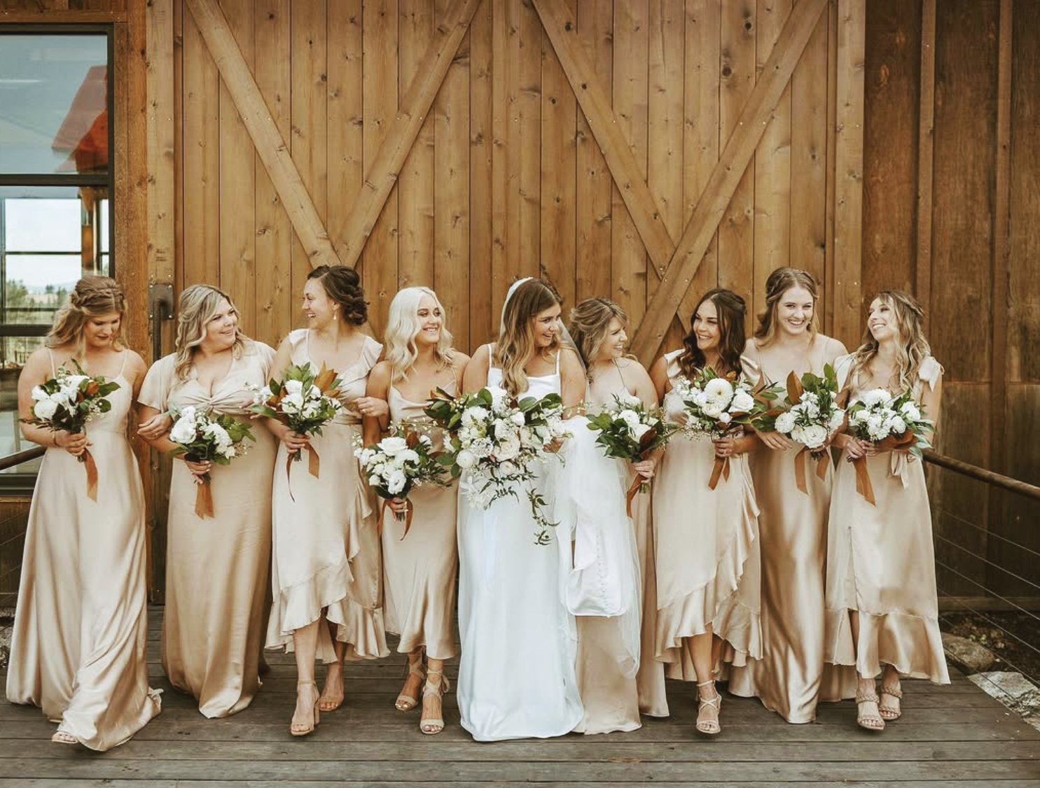 A bride and eight bridesmaids standing in a row, holding bouquets, in front of a wooden barn door during a wedding.