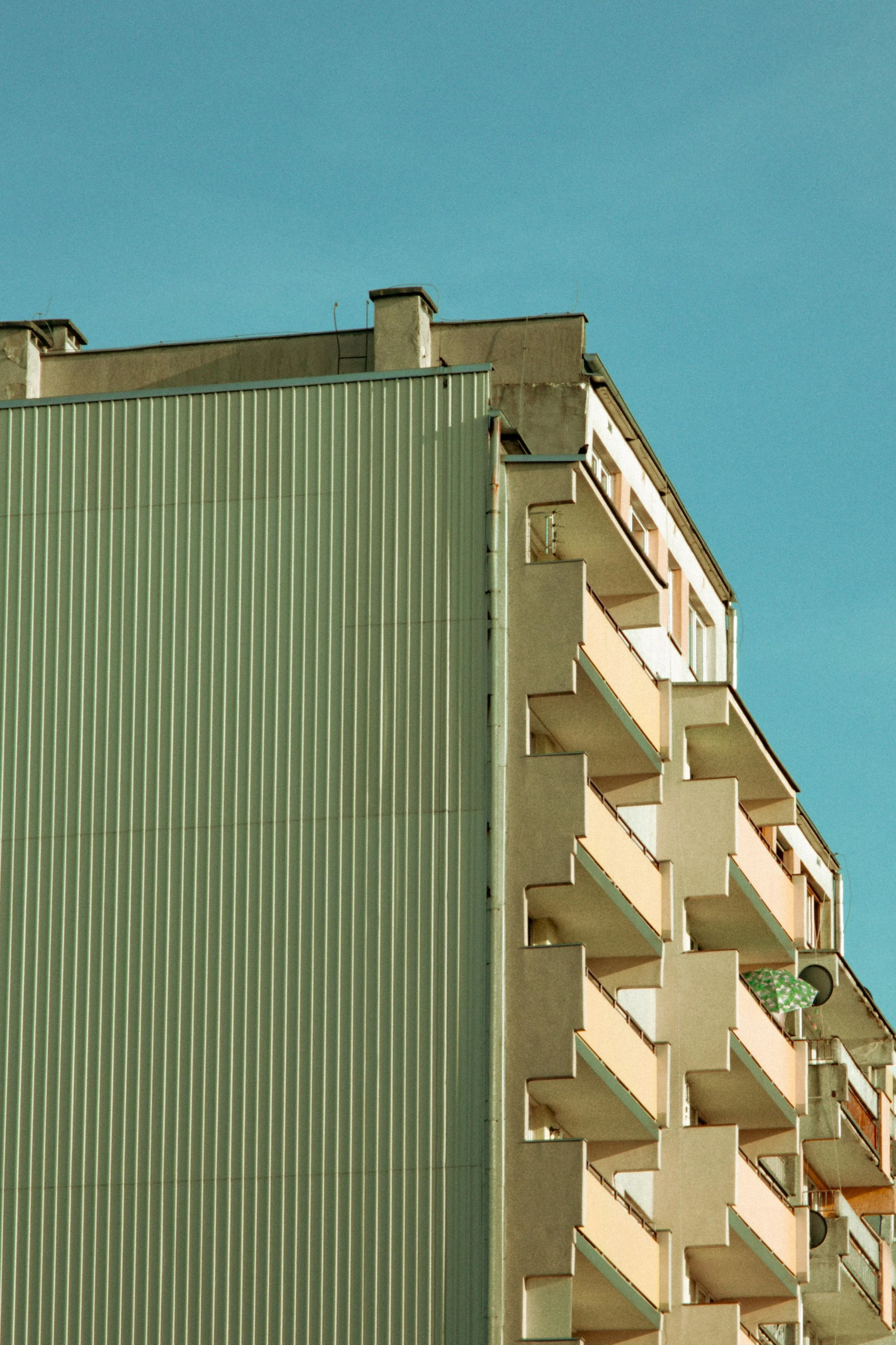 Side view of a multistory residential apartment building with balconies, and a separate metallic facade, against a clear blue sky.