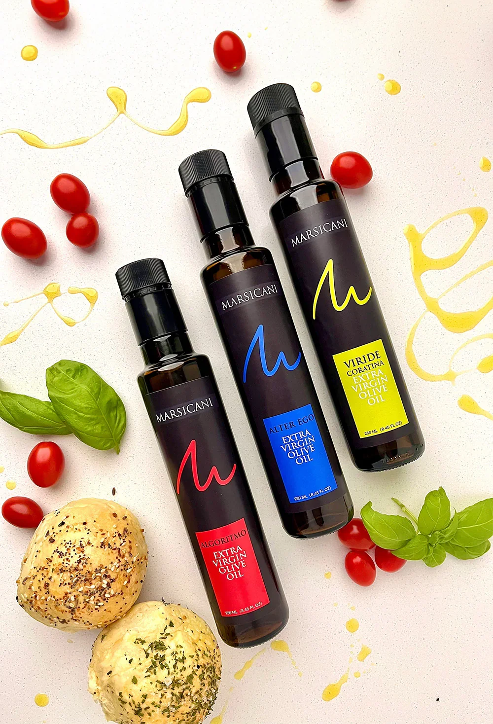 Three bottles of EVOO centered around cherry tomatoes, spinach and bread rolls