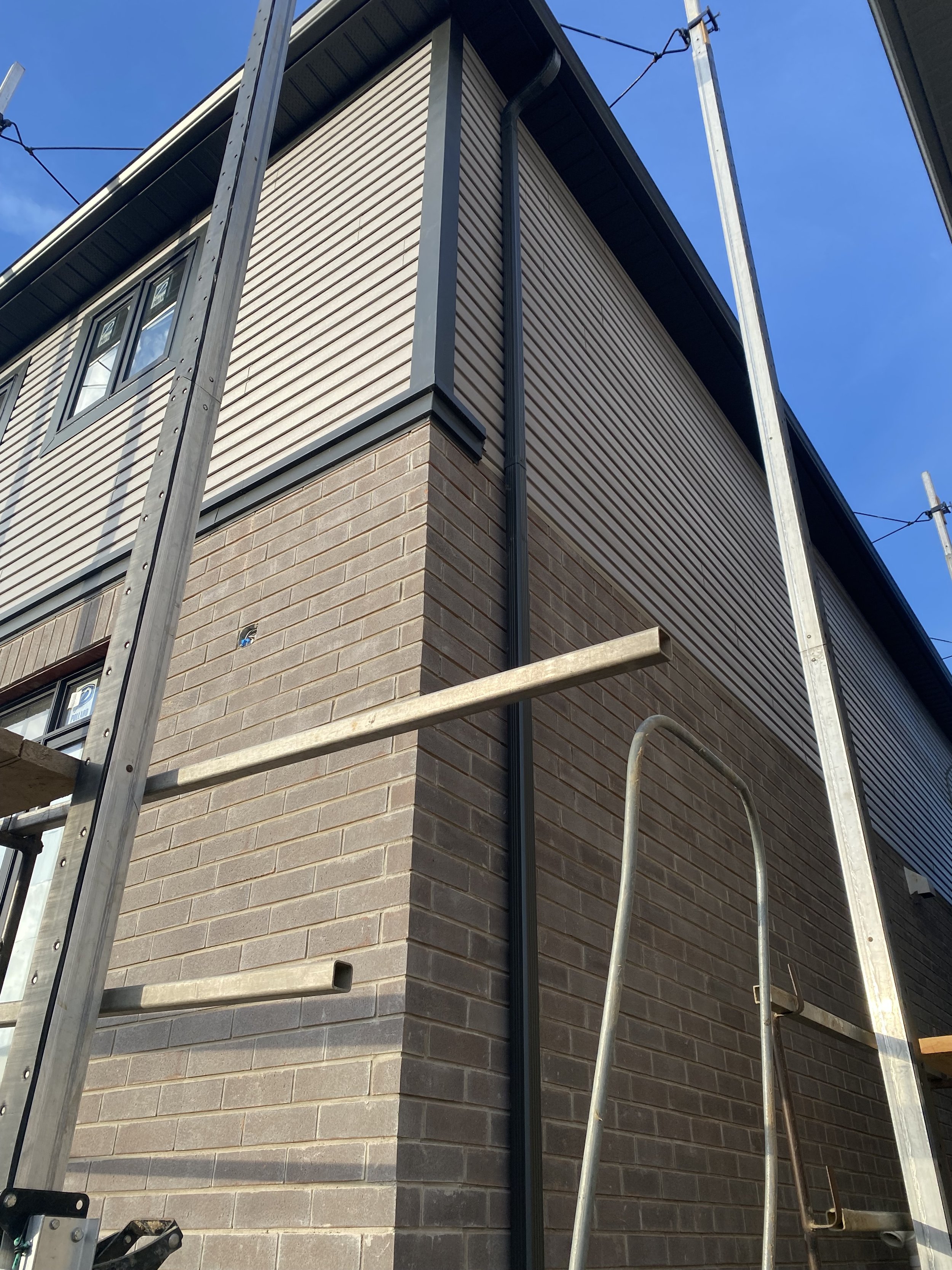 Close-up of a corner of a building under construction, showing brickwork, siding, and scaffolding with ladders, against a blue sky.