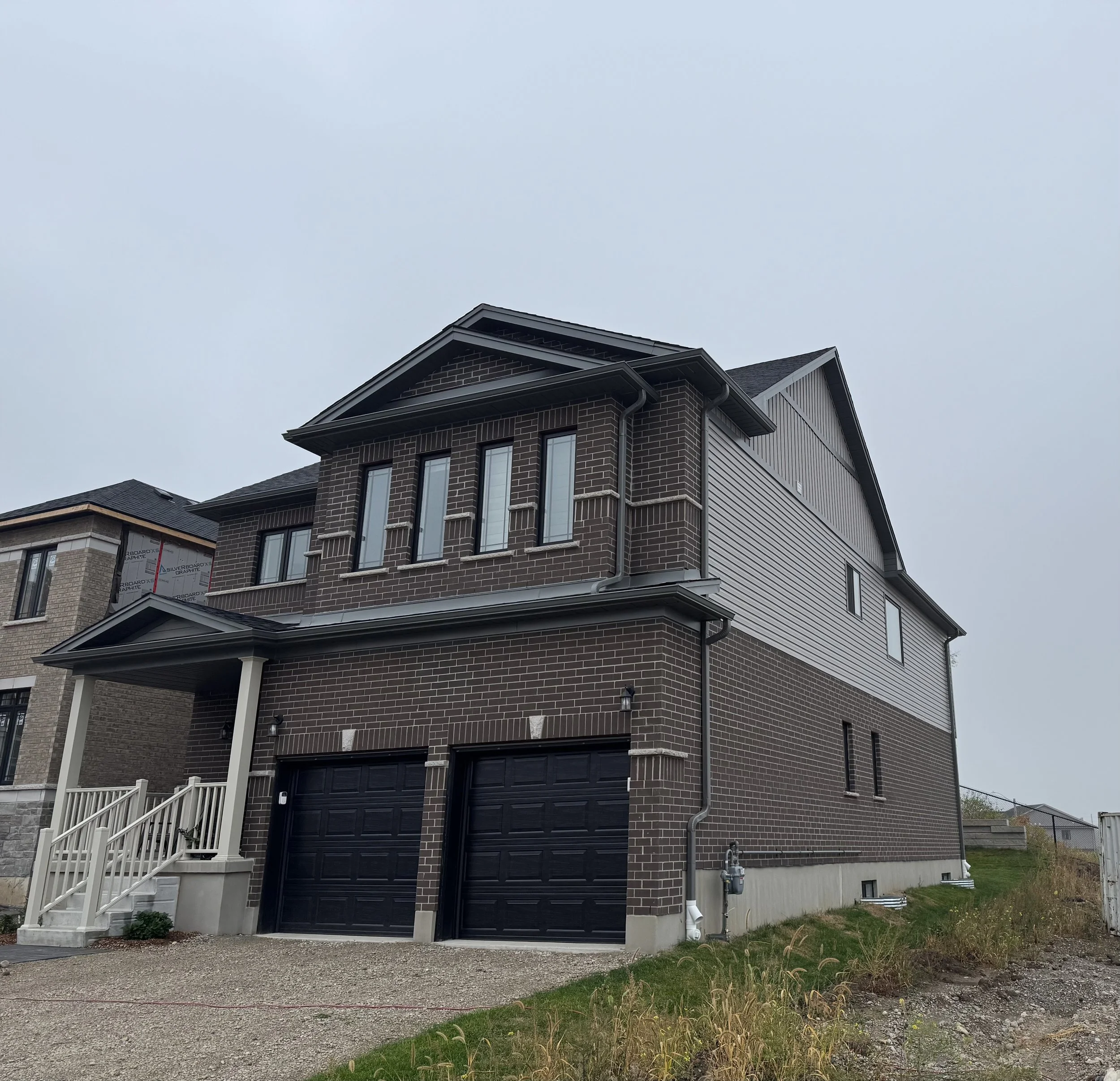 A modern, multi-story brick house with a black garage door, small front porch with stairs, and a driveway, under an overcast sky.