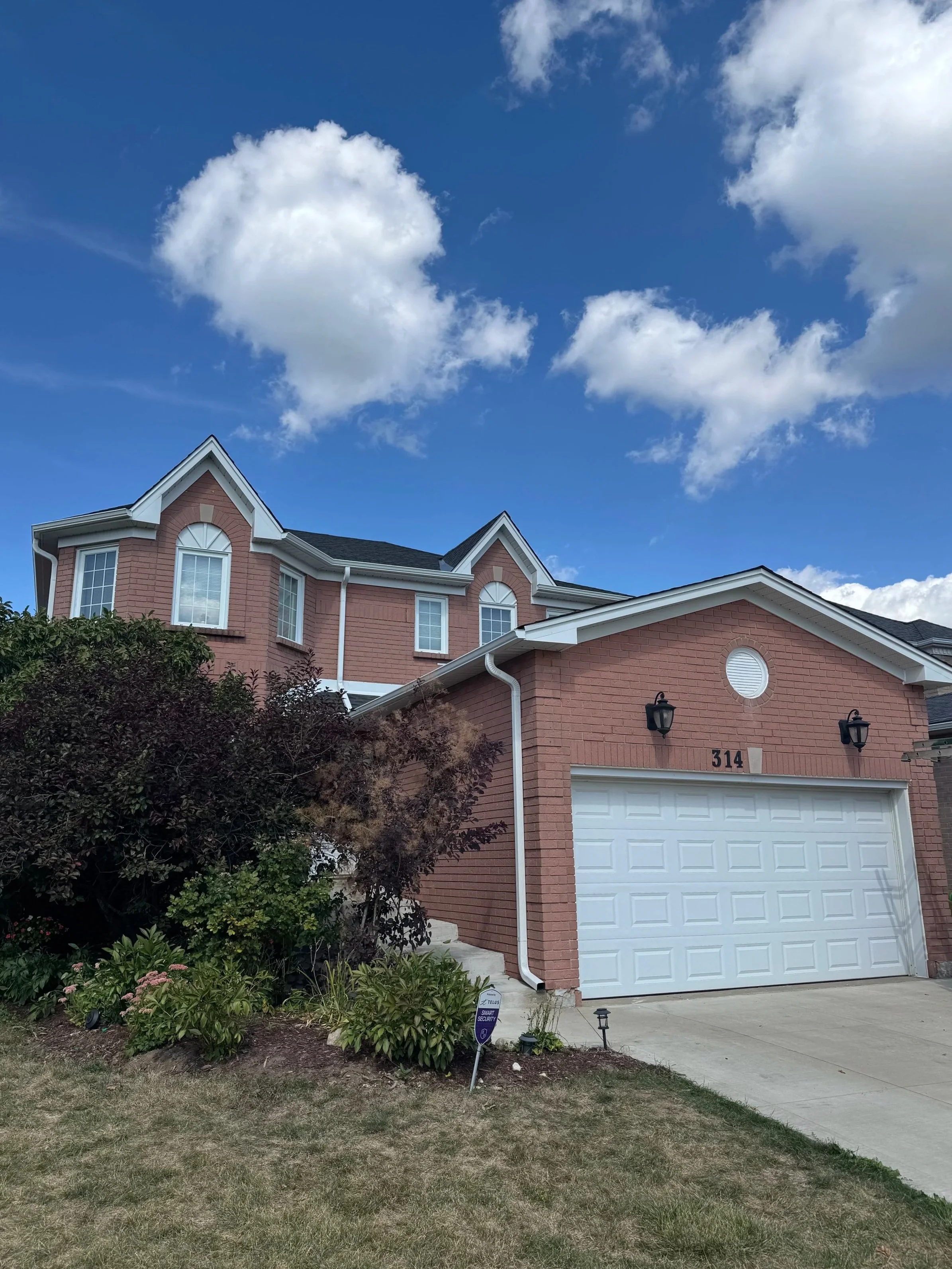 A two-story brick house with white trim and a white garage door, number 314, under a partly cloudy blue sky, with a small front yard and landscaped bushes.