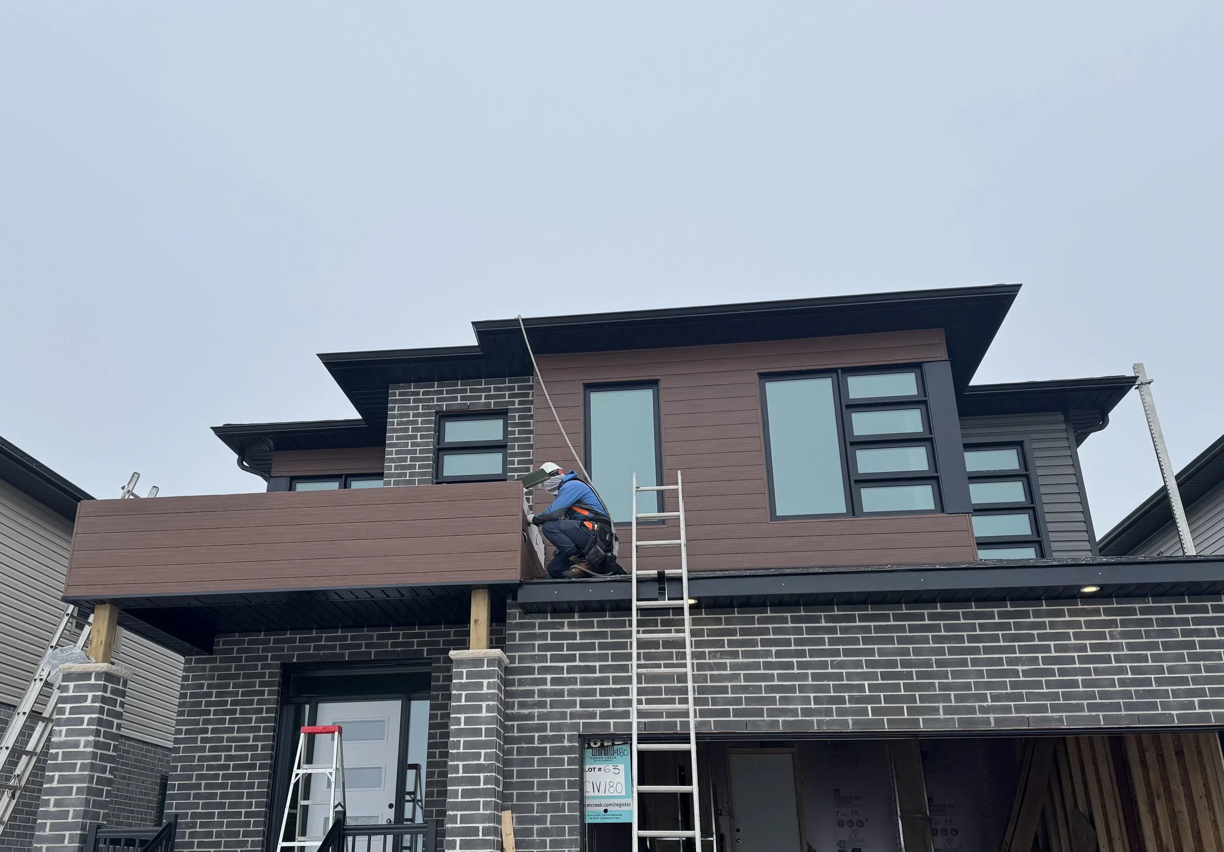 A construction worker installing siding on a modern two-story house with a brick and wood exterior, ladders, and scaffolding.