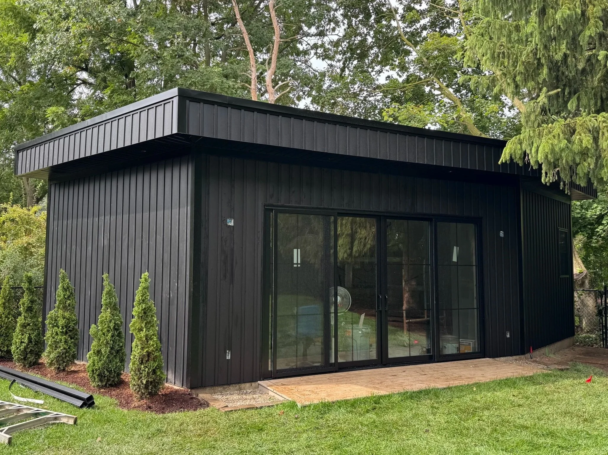 Black modern tiny house with sliding glass doors and a small wooden porch, surrounded by green grass and trees.