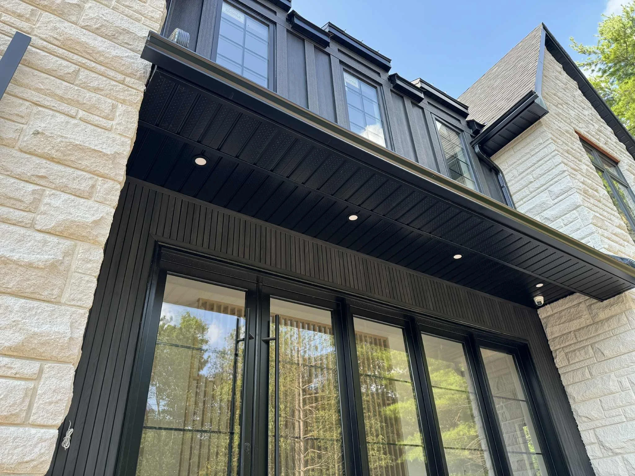 Close-up of the modern house exterior featuring black metal and stone siding, large glass doors, and a balcony with a black metal railing and trimmings, with trees and a blue sky reflected in the glass.