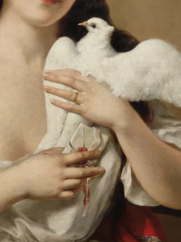 A person holding a white dove close to their chest, with fingers gently gripping the bird's body and wing. The person is wearing a ring on their left hand, and the background is neutral beige.