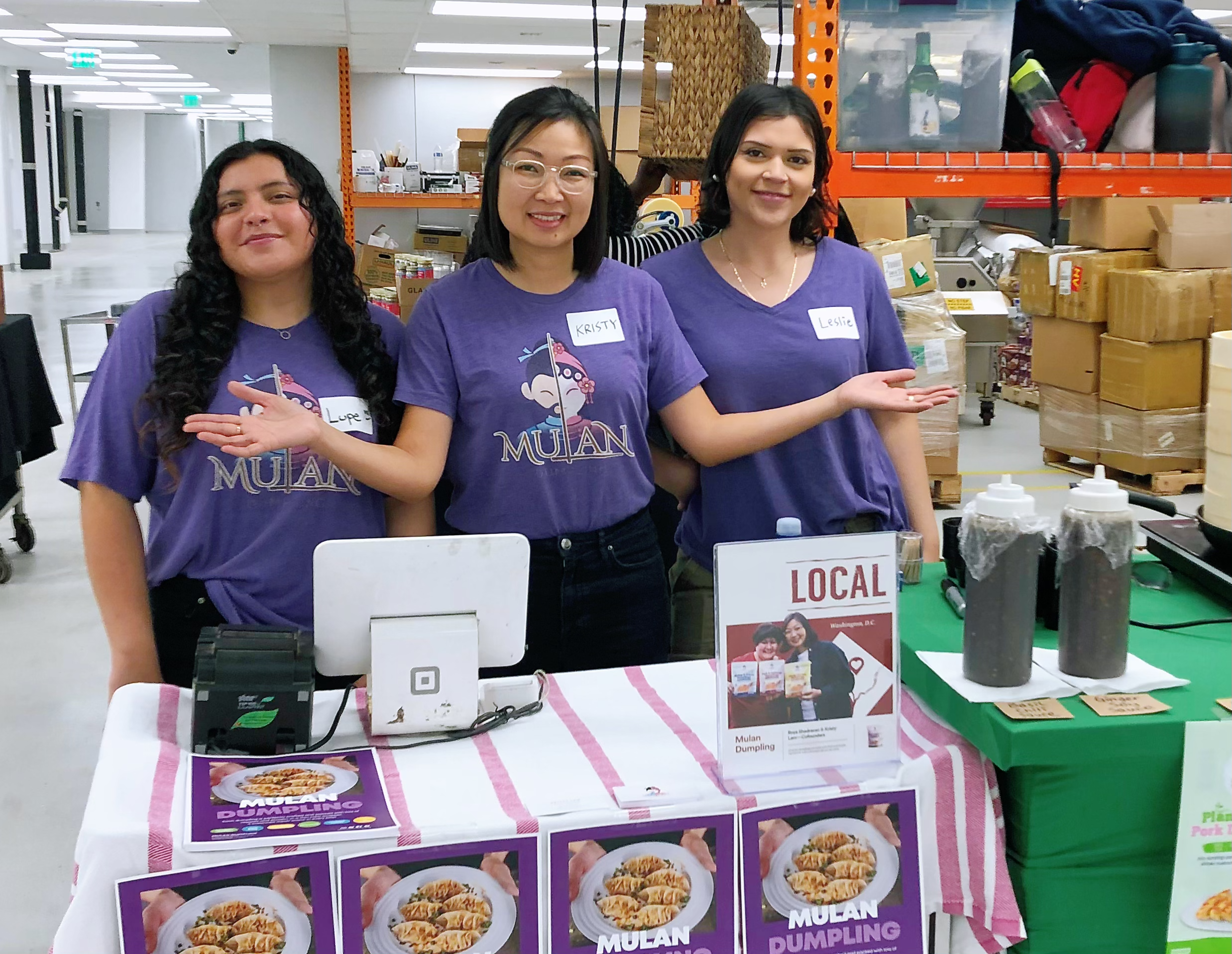 Three women standing behind a table at a food stall, all wearing purple T-shirts with 'Mulan' printed on them. The woman in the middle has a name tag that says 'Kristy.' The table displays promotional flyers and pictures of dumplings, with containers of dark sauces or condiments on the right. The background shows a warehouse or storage area with boxes and shelving.