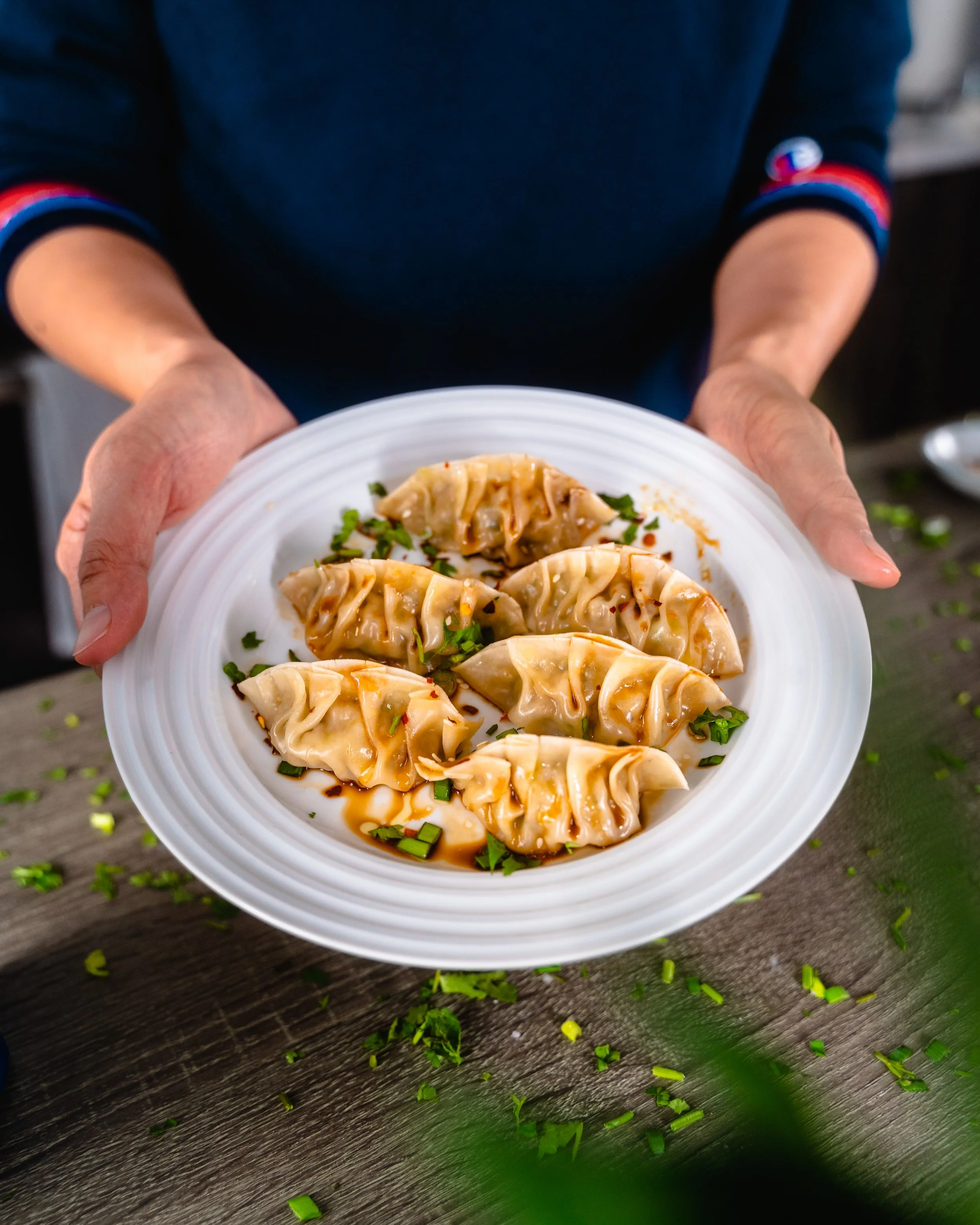 Person holding a white plate with five cooked dumplings garnished with chopped green herbs and sauce.