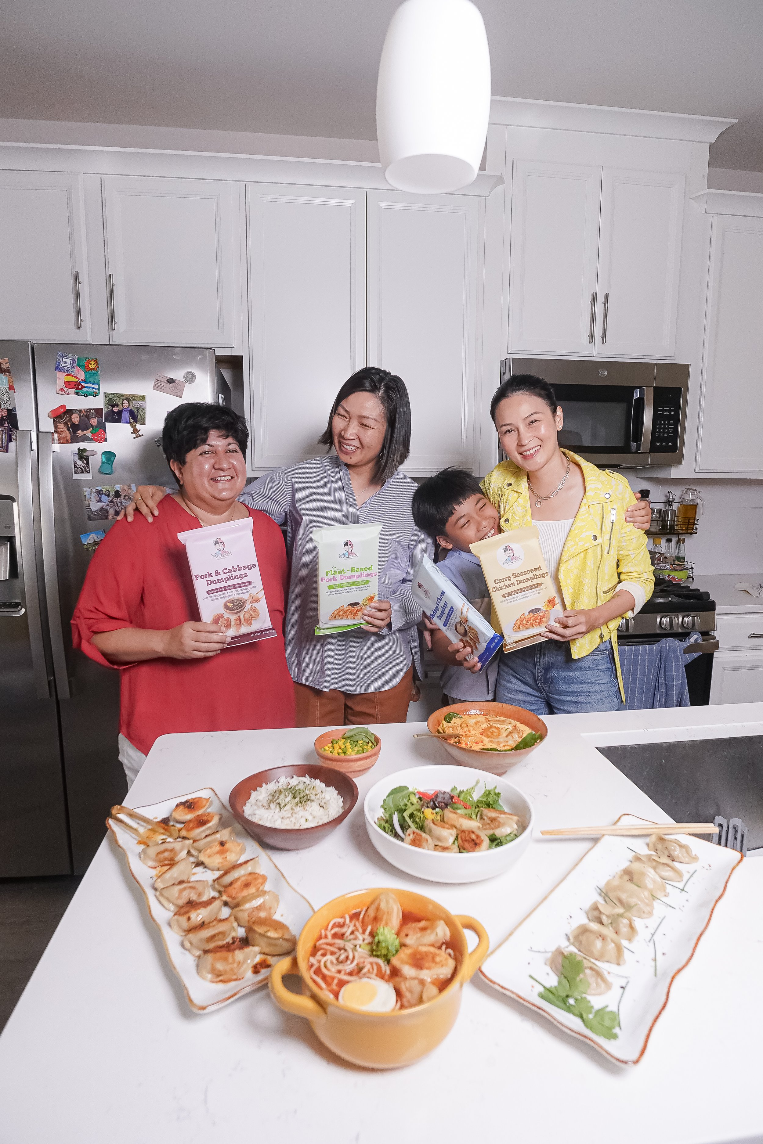 Group of four people in a kitchen smiling and holding Asian food recipe booklets, with various Asian dishes on the counter in front of them.