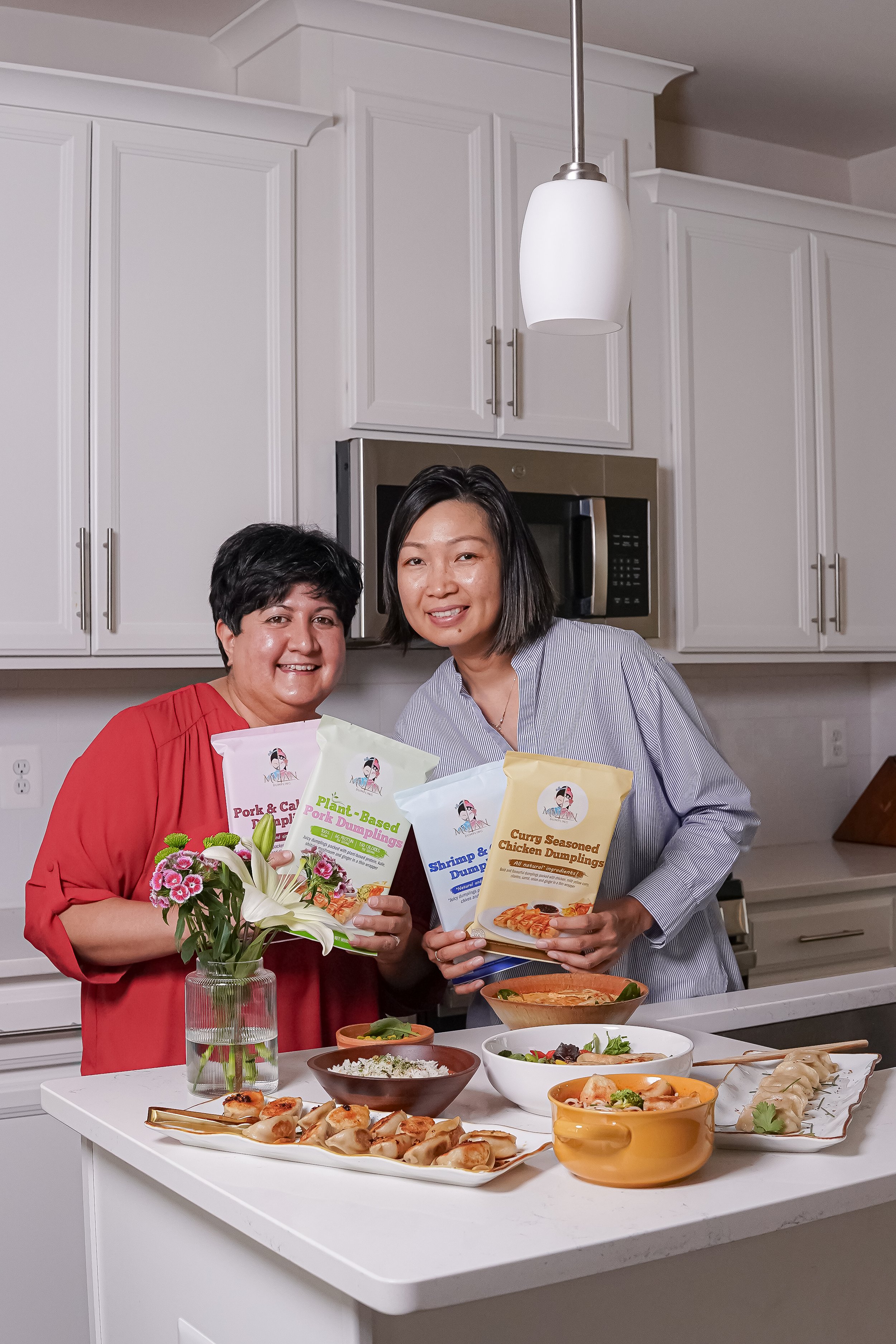 Two women in a kitchen holding packages of plant-based dumplings, surrounded by bowls of food on a white kitchen island with white cabinets in the background.