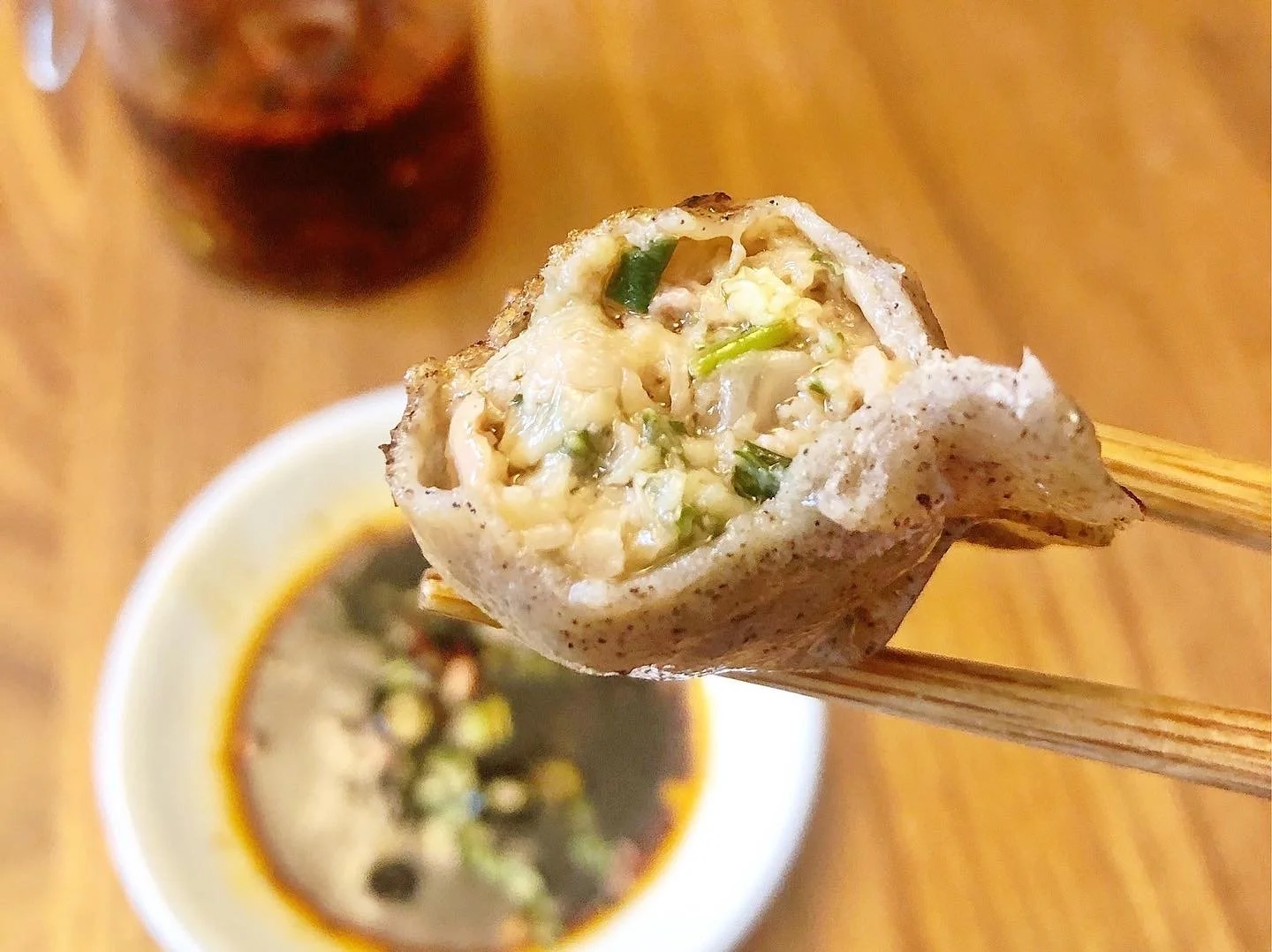 Close-up of a chopstick holding a piece of Vietnamese spring roll filled with chopped vegetables and shrimp, with dipping sauce and a drink in the background.