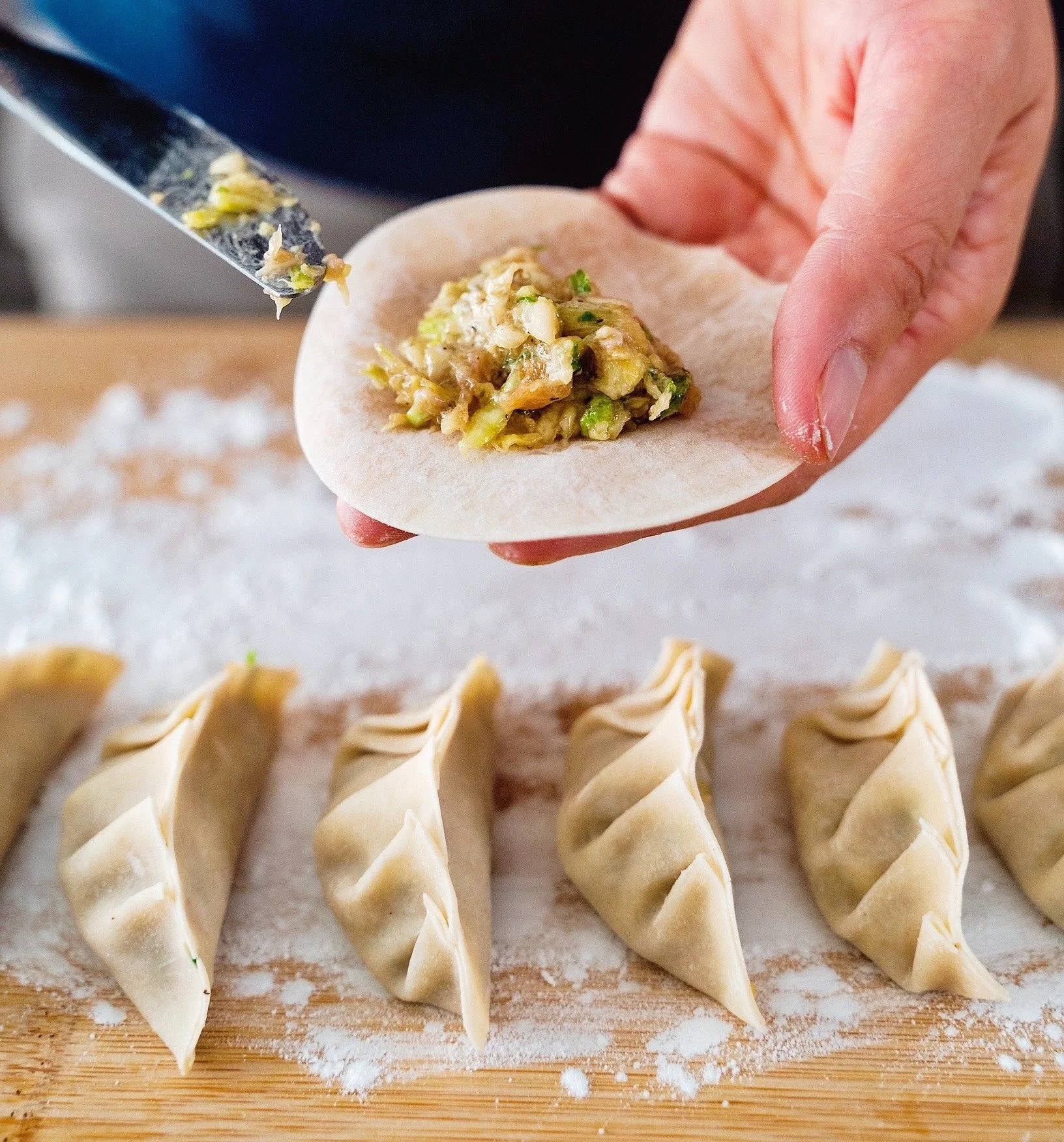 Close-up of a person filling dumplings with a mixture of vegetables and meat on a wooden surface sprinkled with flour, with assembled dumplings lined up in the foreground.
