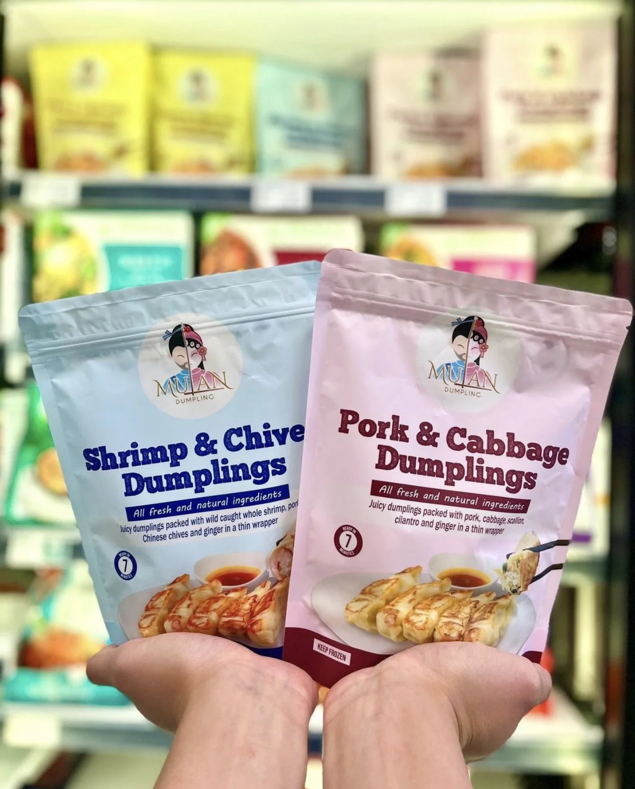 Two hands holding packages of frozen Mulan dumplings, one shrimp and chive flavor in a blue package, and one pork and cabbage flavor in a pink package, in a grocery store aisle.