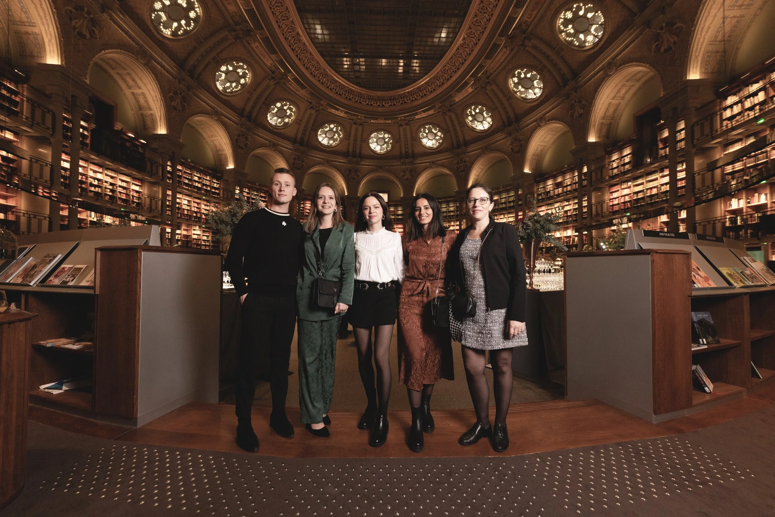Cinq personnes debout dans une bibliothèque ornée de livres, avec une architecture en dôme et de grandes fenêtres rondes.