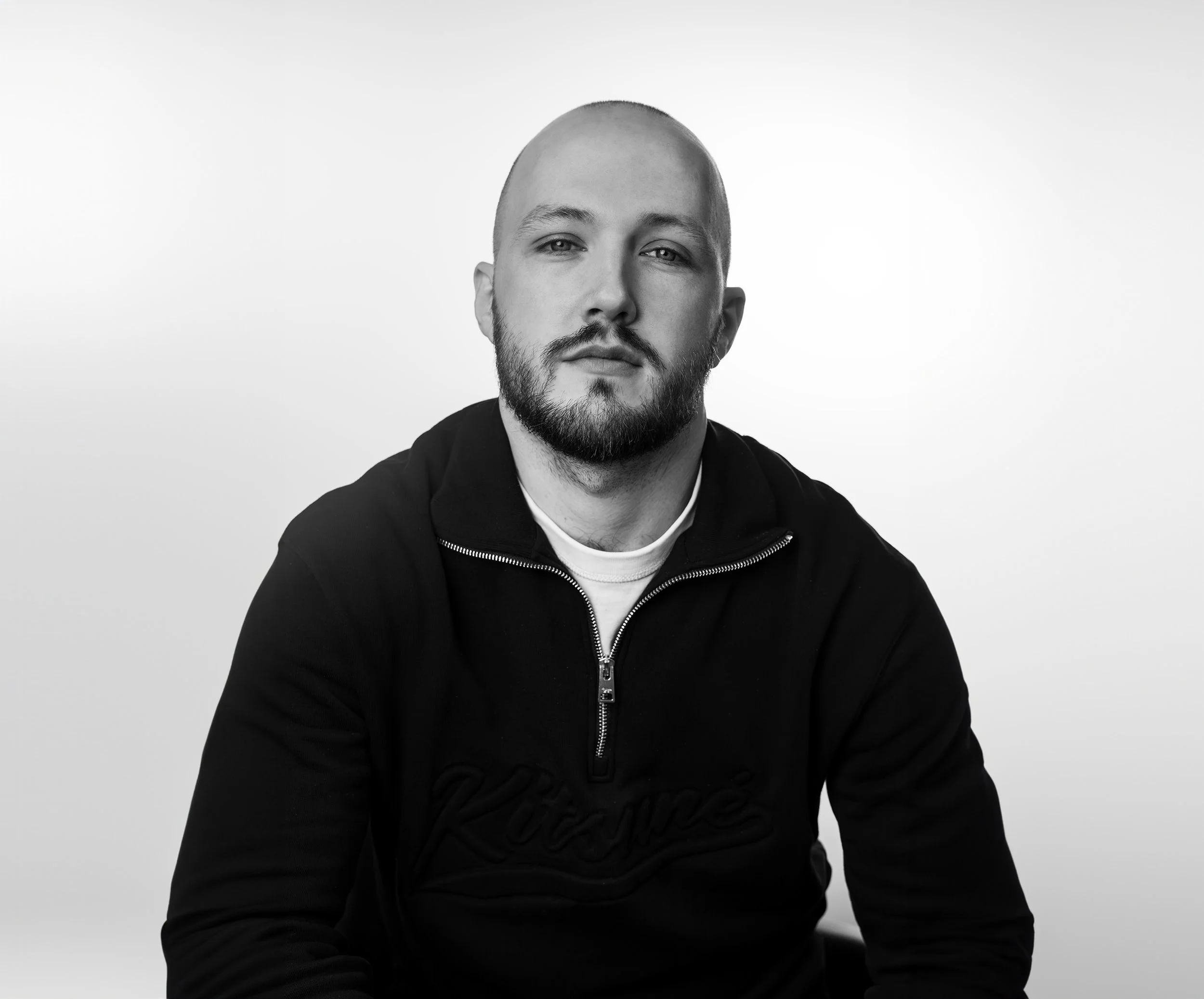 Un homme avec une barbe, assis contre un fond blanc, portant un sweat à demi-zippé