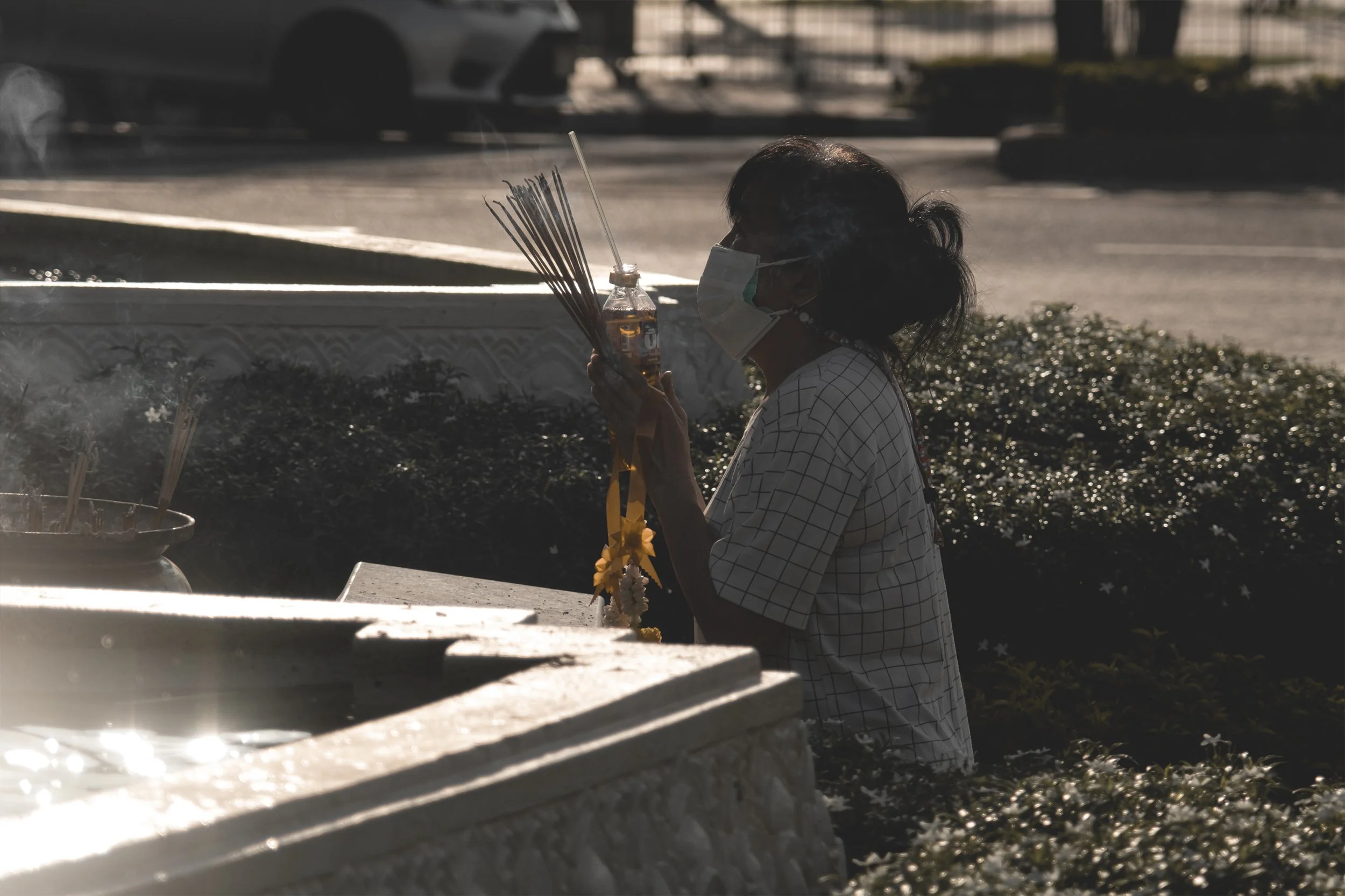 Une femme portant un masque facial, tenant un breuvage et des bâtons d'encens, en train de prier ou de faire une offrande à l'extérieur, silhouette sombre contre la lumière du jour.
