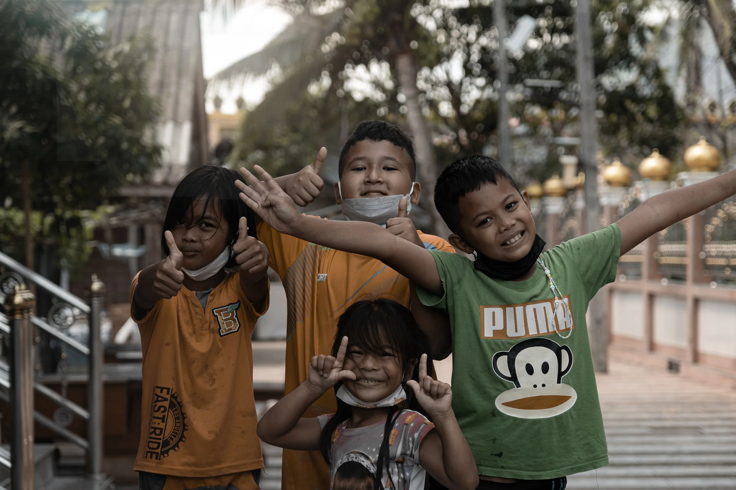 Groupe de quatre enfants souriants et faisant des gestes de pouces en l'air, avec des masques faciaux abaissés ou enlevés, à l'extérieur, dans un environnement arboré et lumineux.